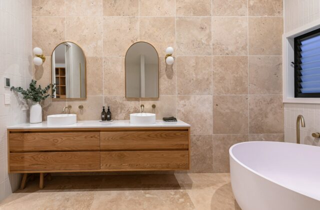 Travertine bathroom with a timber vanity, twin basins and brass tapware