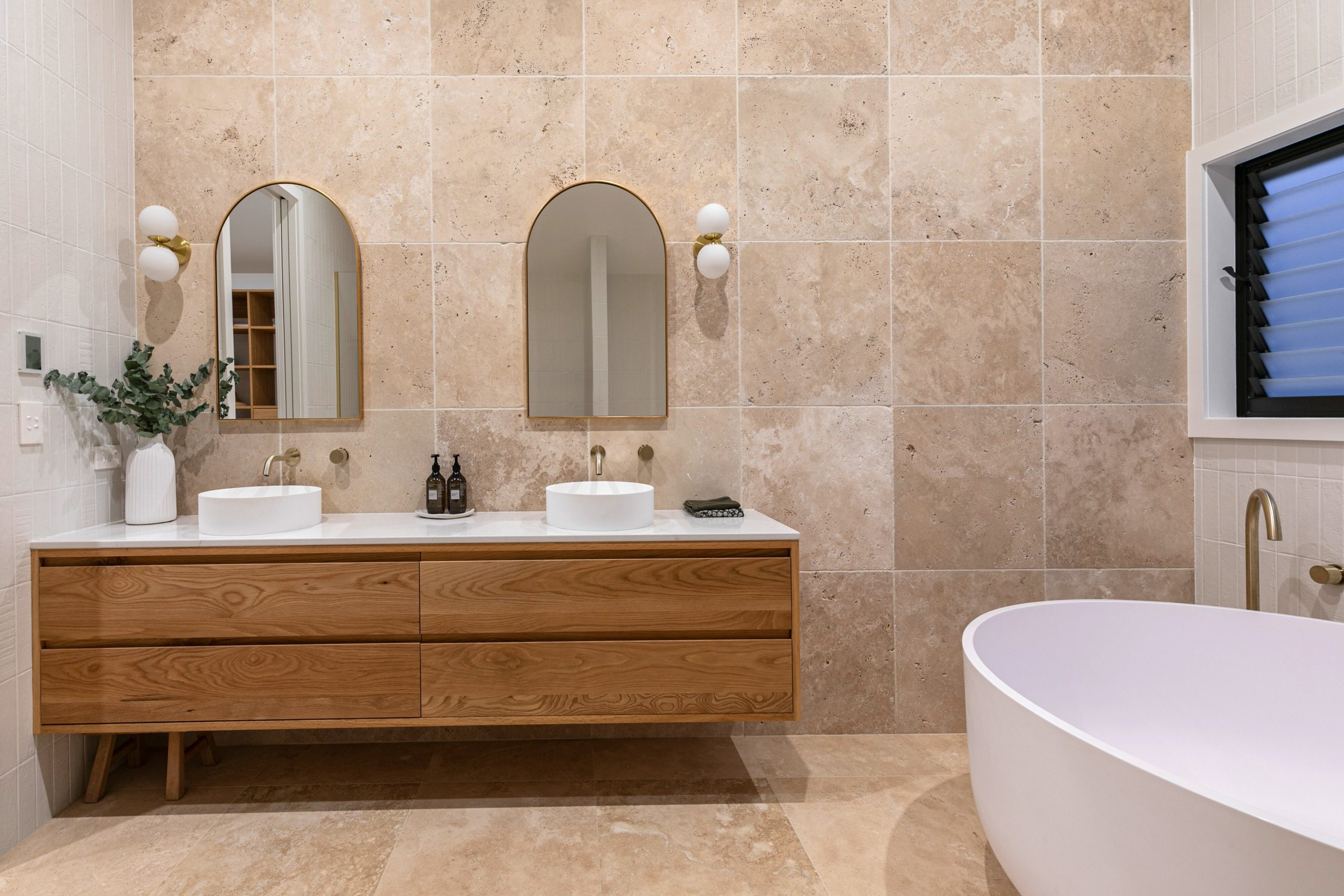 Travertine-tiled bathroom with a double oak vanity, arched mirrors and a freestanding bath